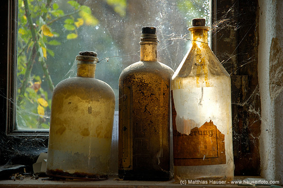 Drei alte verstaubte Glasflaschen mit Spinnweben vor einem Fenster | Three old and dusty glass bottles