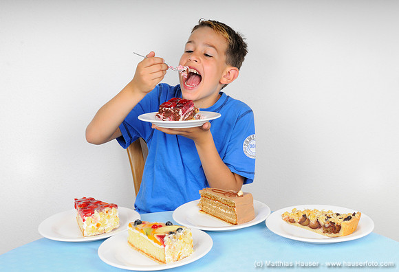 Junge isst Kuchen, auf einem Tisch stehen vier weitere Teller mit Kuchenstücken - Boy eating cake at a table with four plates with other pieces of cake